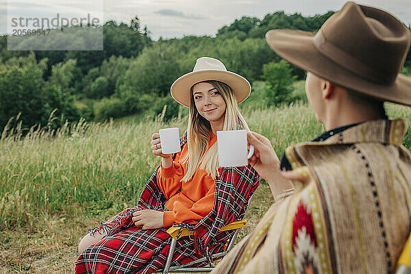 Smiling woman drinking tea and discussing with boyfriend at meadow on mountain