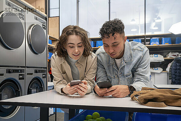 Young woman and man using mobile phones leaning on table at laundromat