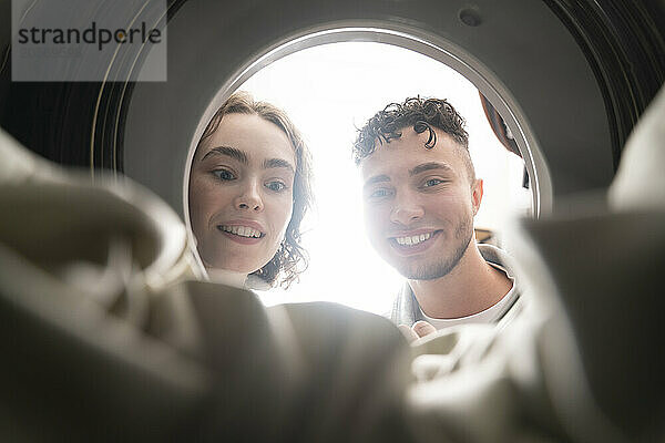 Smiling young couple looking inside of washing machine at laundromat