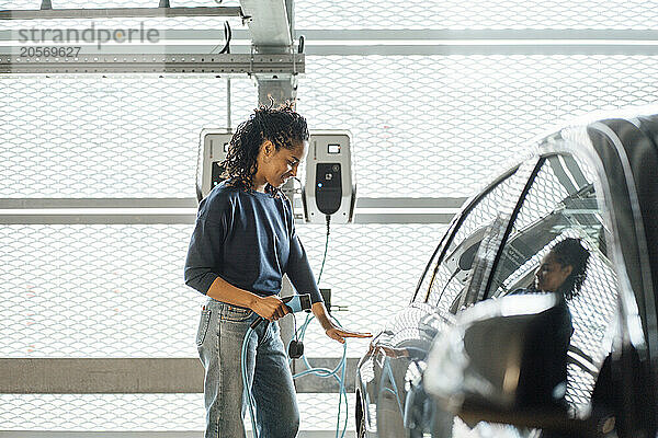 Smiling young businesswoman charging electric car with plug at parking garage