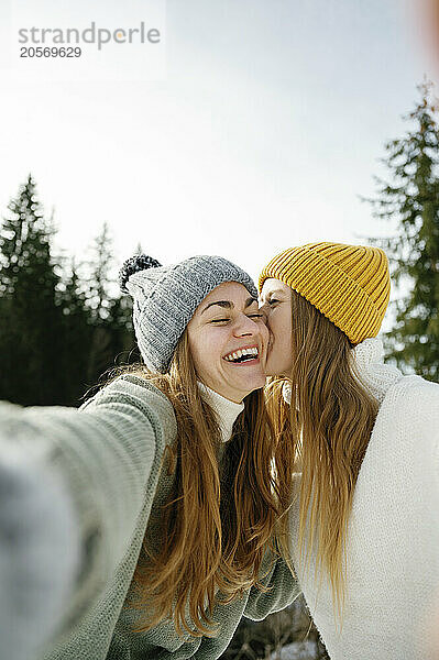 Woman kissing cheerful friend taking selfie