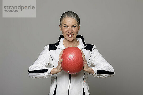 Smiling active senior woman holding red ball against gray background