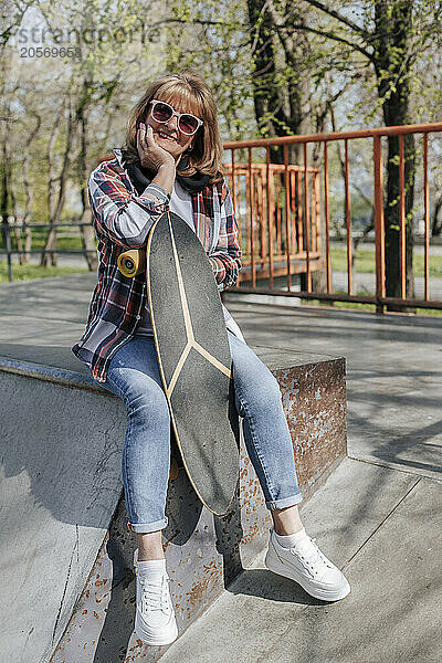 Smiling senior woman leaning on skateboard and sitting at park