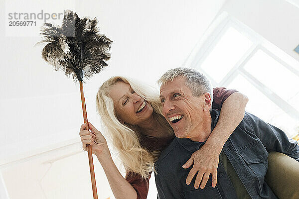 Happy senior man piggybacking blond woman with duster at home