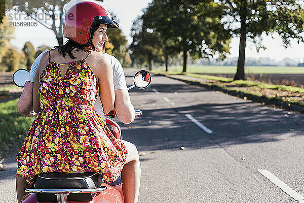 Smiling young woman wearing helmet and sitting on motor scooter with boyfriend at country road