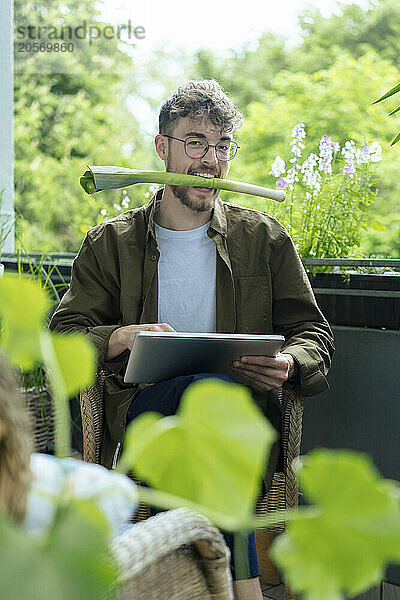 Playful young man with leek in mouth sitting at balcony