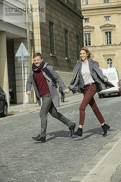 Happy boyfriend and girlfriend holding hands and crossing street in city