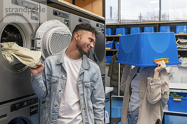 Smiling young man looking at playful girlfriend covering face with blue basket in laundromat