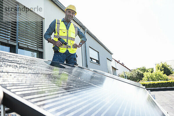 Confident mature technician with power tool by solar panel at rooftop