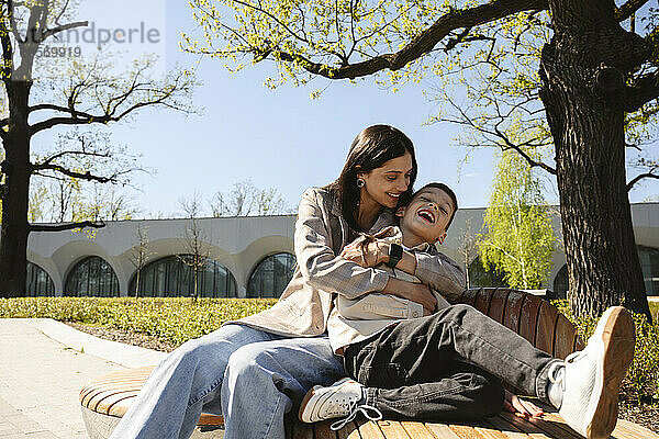 Mother embracing son and sitting on bench in park