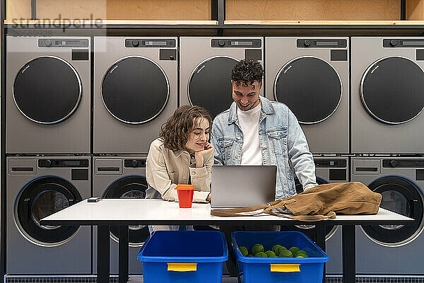 Smiling young couple using laptop on table in front of washing machines waiting at laundromat