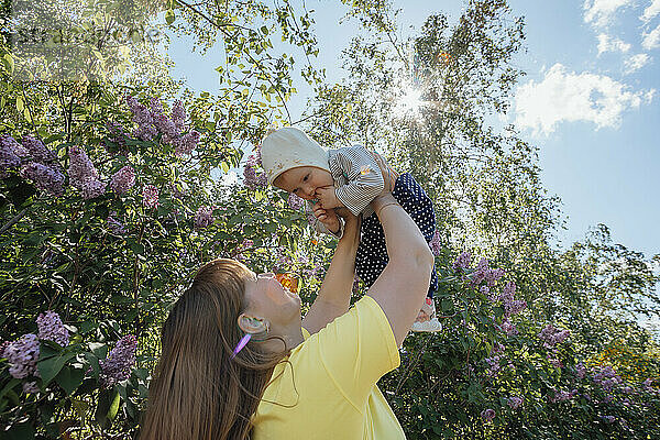 Happy mother playing and carrying baby girl near flower tree in garden