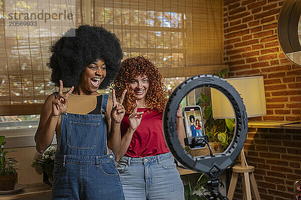 Happy multiracial friends doing peace sign and vlogging through smart phone at home