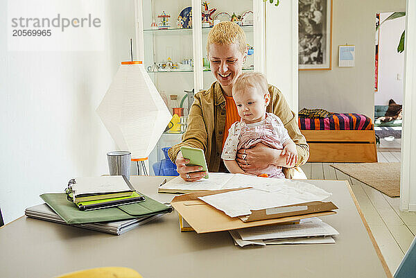 Smiling mother sitting with toddler daughter and using smart phone at home