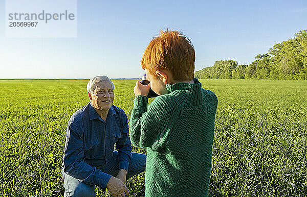 Boy photographing grandfather on camera in field