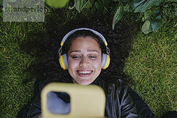 Happy young woman using smart phone and listening to music through wireless headphones lying on grass at park