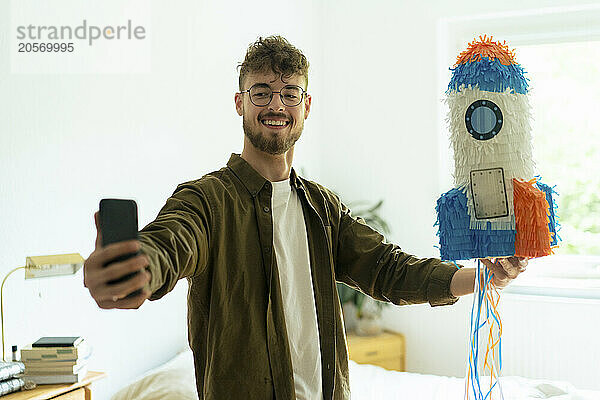 Young man taking selfie with rocketship shaped pinata lying on bed at home