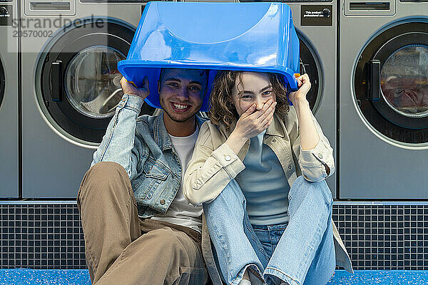 Playful young couple with blue basket over head sitting in front of washing machines at laundromat