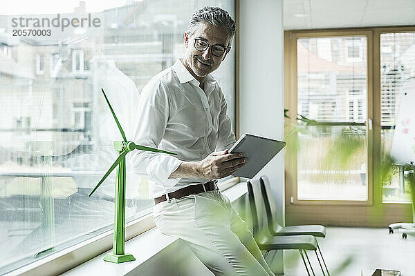 Mature businessman with tablet PC looking at wind turbine model in office