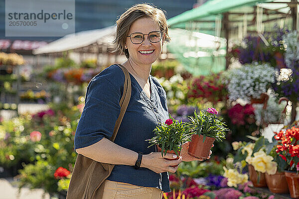 Smiling woman in eyeglasses holding flower pot and standing in nursery