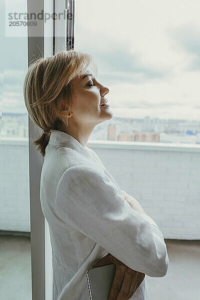 Smiling mature businesswoman with eyes closed leaning on glass window