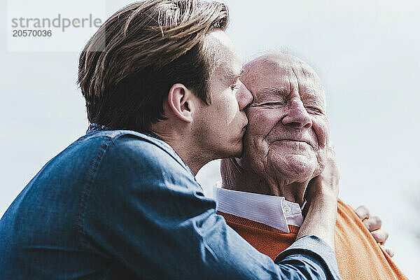 Young man kissing happy grandfather under sky