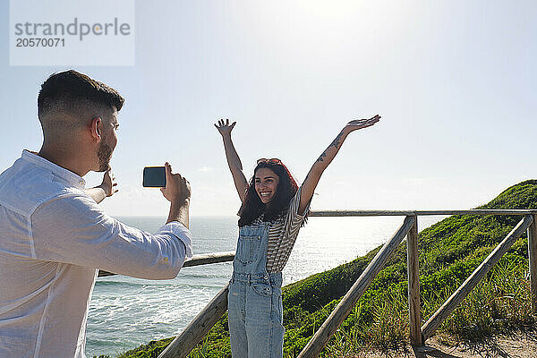 Man photographing girlfriend standing with arms raised near railing