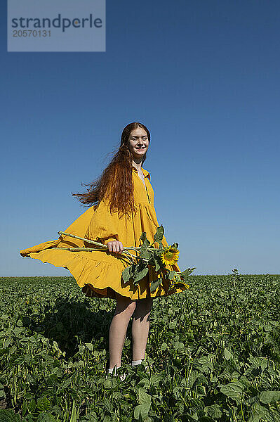 Teenage girl with red hair in a yellow dress holding sunflowers in a green field