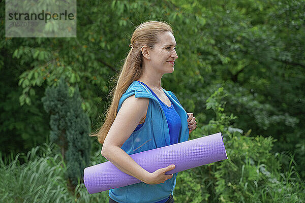 Smiling woman holding yoga mat and walking at park