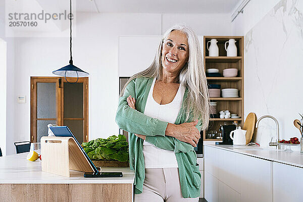 Happy mature woman standing with arms crossed in kitchen at home