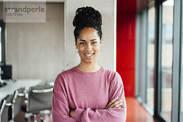 Happy young businesswoman standing with arms crossed in front of column at office