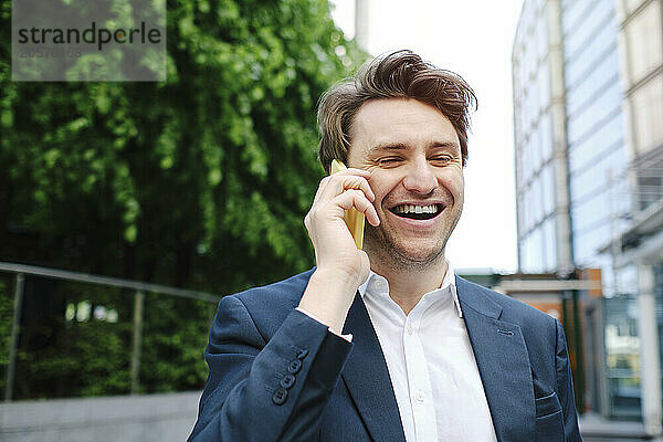 Cheerful businessman talking on smart phone in front of trees and building