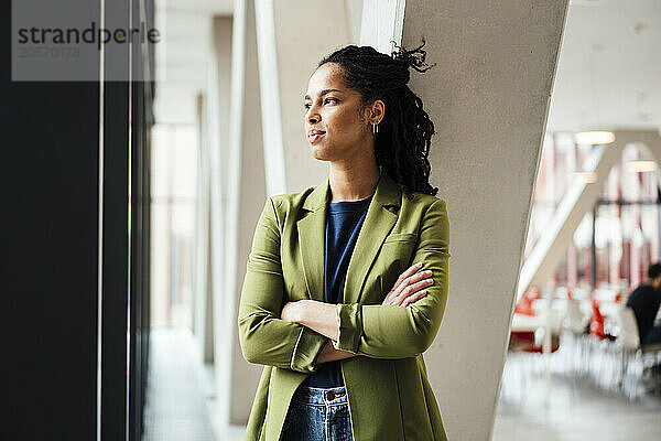 Thoughtful young businesswoman standing with arms crossed looking out through window at office