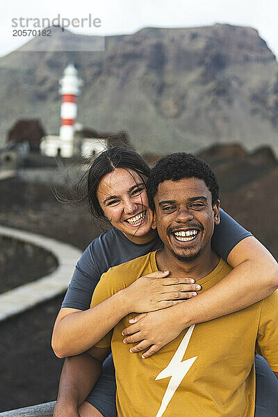 Happy young woman with arm around boyfriend in front of Punta Teno Lighthouse