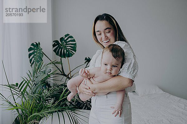 Happy mother playing with baby boy near plants at home