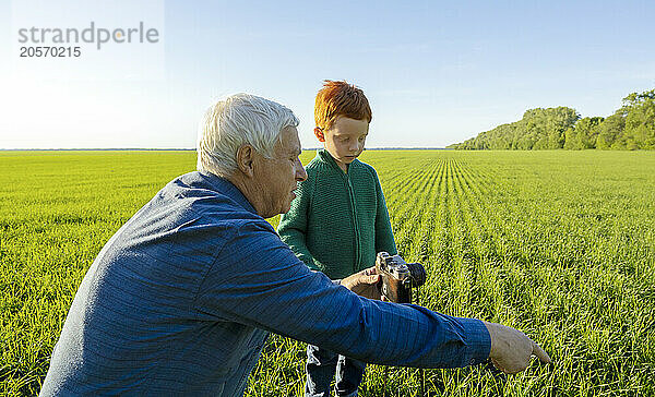 Senior man teaches grandson how to take photograph in field