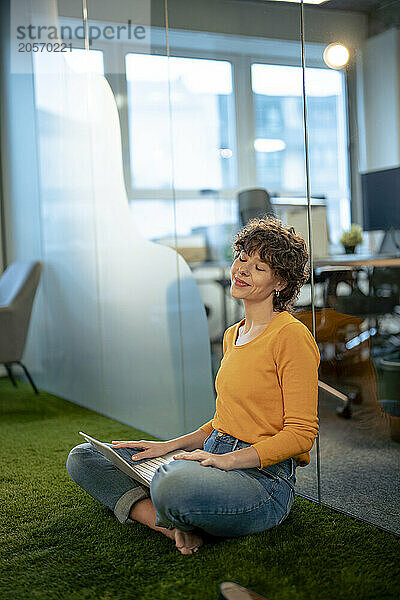 Smiling businesswoman sitting with eyes closed and laptop near glass in office