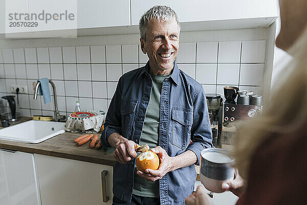 Happy man peeling orange looking at woman in kitchen