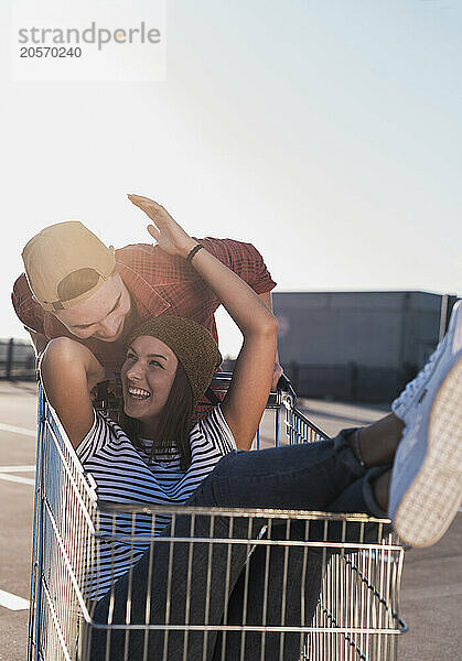 Playful young woman sitting in shopping cart and enjoying with boyfriend under clear sky