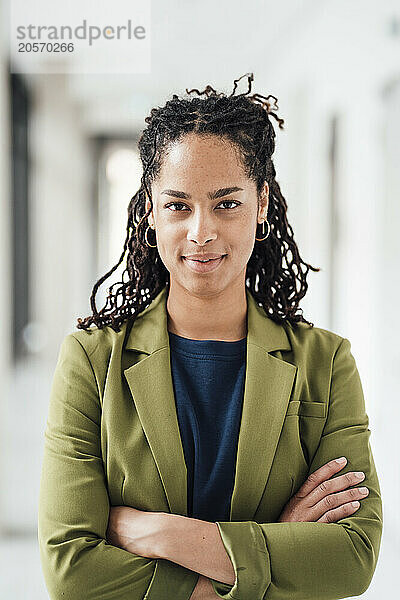 Confident young businesswoman with arms crossed standing in office corridor