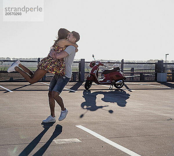 Happy young man embracing girlfriend and standing at parking lot under clear sky