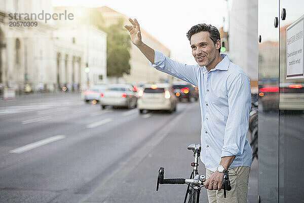 Handsome mature businessman with bicycle waving at sidewalk in city