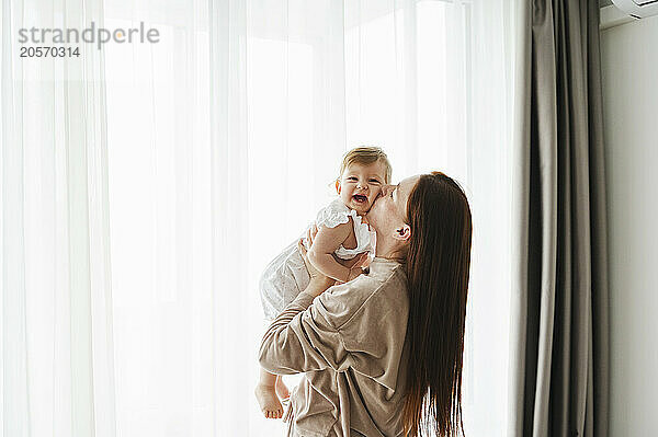 Woman kissing daughter near window at home