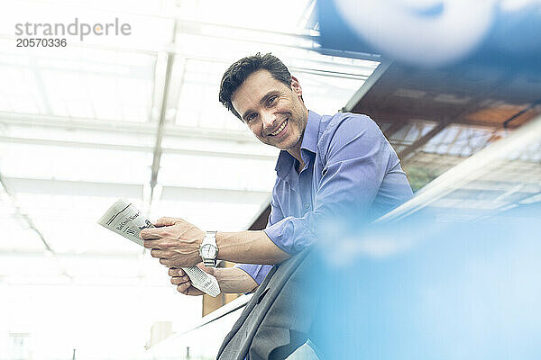 Smiling mature businessman with newspaper leaning on railing at airport terminal