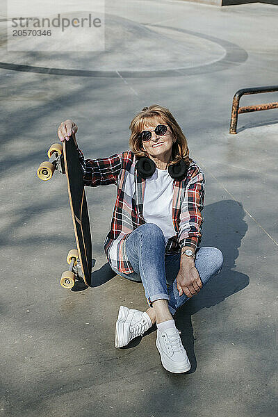 Happy senior woman sitting with skateboard on sports ramp in park