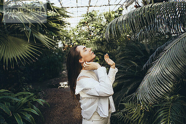 Young woman with eyes closed standing near trees in greenhouse