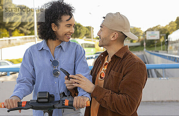 Smiling woman holding electric push scooter and standing with boyfriend at park