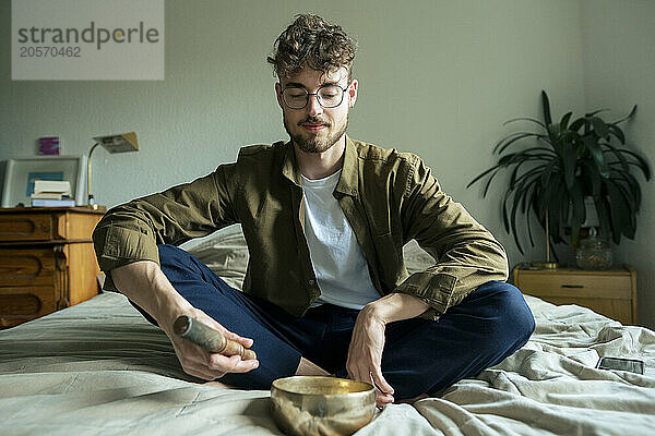 Handsome young man meditating with singing bowl on bed at home