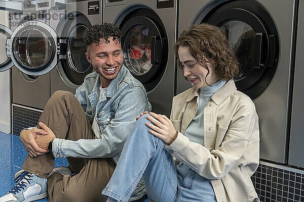 Smiling young friends sitting together leaning on washing machines at laundromat