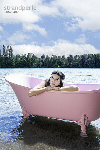 Smiling girl with swimming goggles relaxing inside pink bathtub in lake under cloudy sky